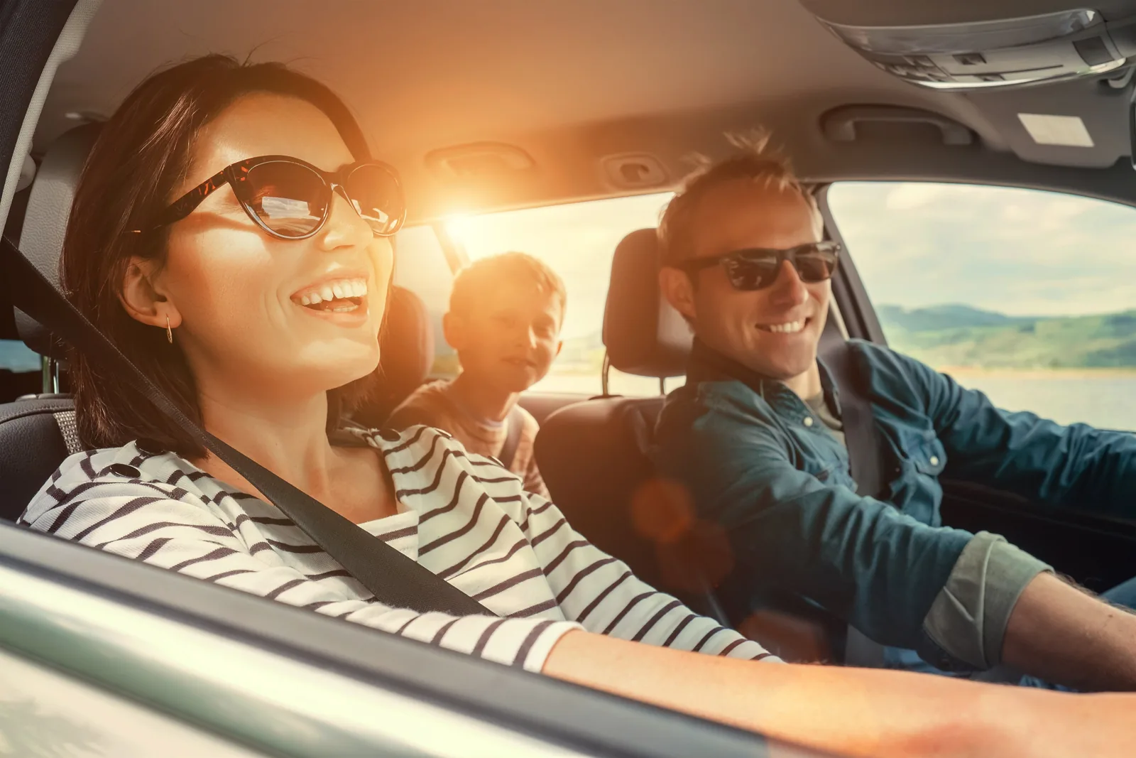 Driver and passenger smiling on a sunny drive with a child in the back seat