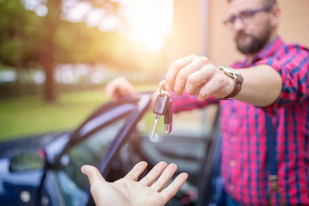 Private seller handing car keys to a buyer in front of a blue hatchback