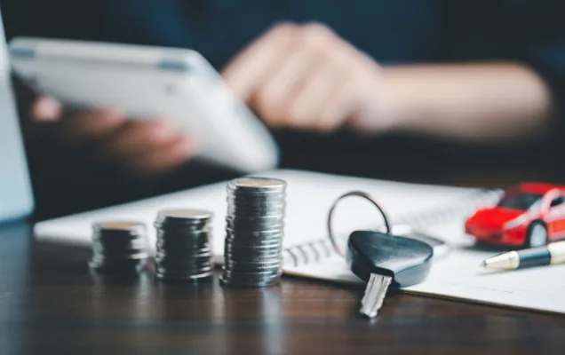 Stacks of coins, a car key and a toy car on a desk beside someone using a tablet
