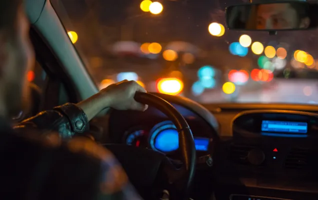 Driver's hand on the wheel at night with city lights blurred beyond the windscreen