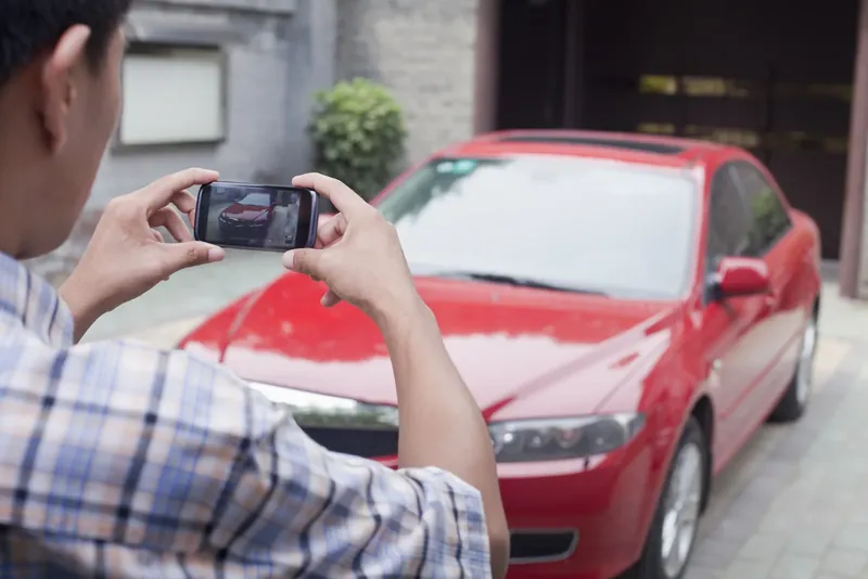 Man photographing a red sedan with his phone in a driveway
