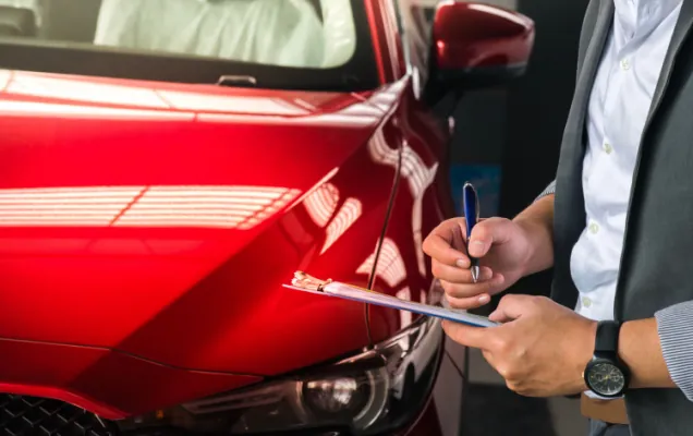 Man filling in transfer paperwork on a clipboard beside a red car in a showroom